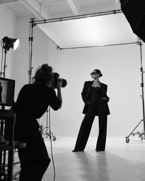 Black and white photographs from a modeling photo shoot: a model in a black suit posing in a studio while a photographer works, and a close portrait in hard window light